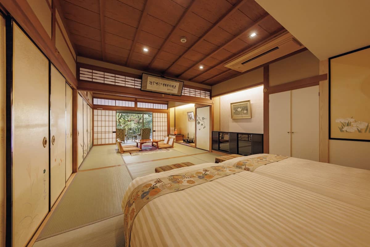A traditional Japanese room with tatami flooring, a bed with a floral bedspread, a television, and a sliding door leading to a view of greenery.