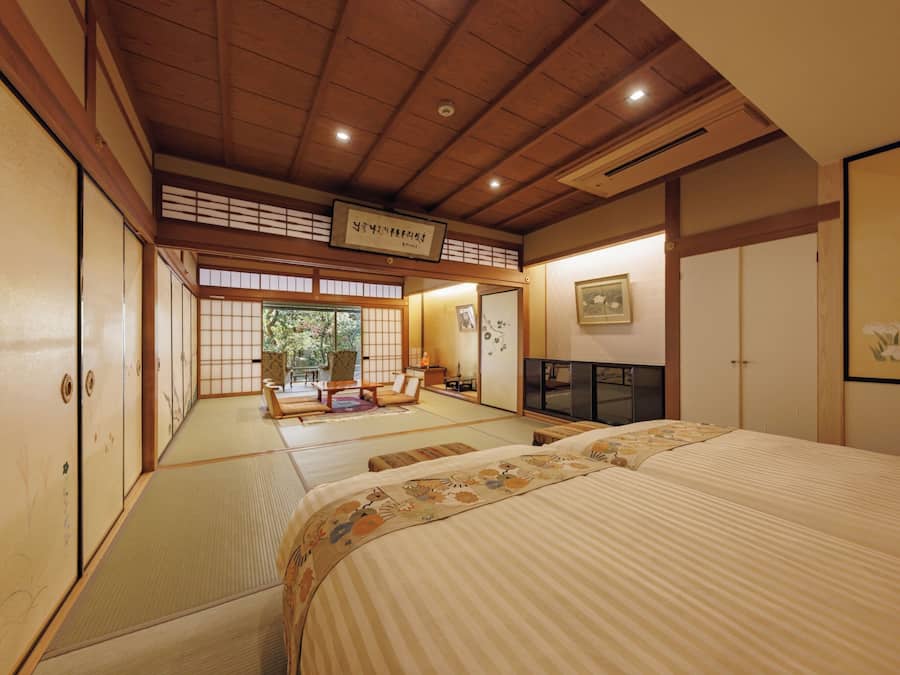 Una habitación tradicional japonesa con suelo de tatami, una cama con cubrecama floral, un televisor y una puerta corrediza que da a vistas de follaje.