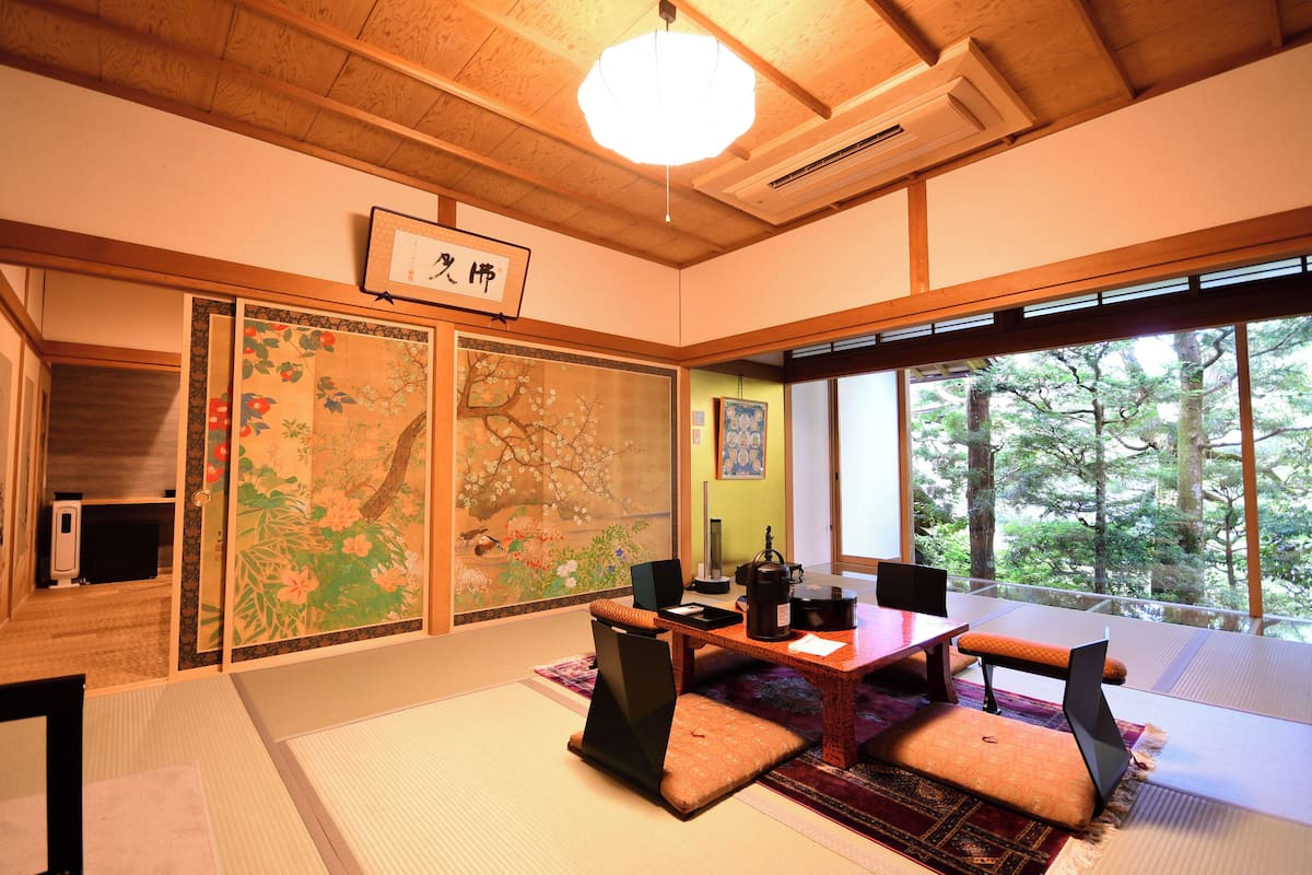 A traditional Japanese room with tatami flooring, a low table with cushions, a television, and a view of trees through large windows.