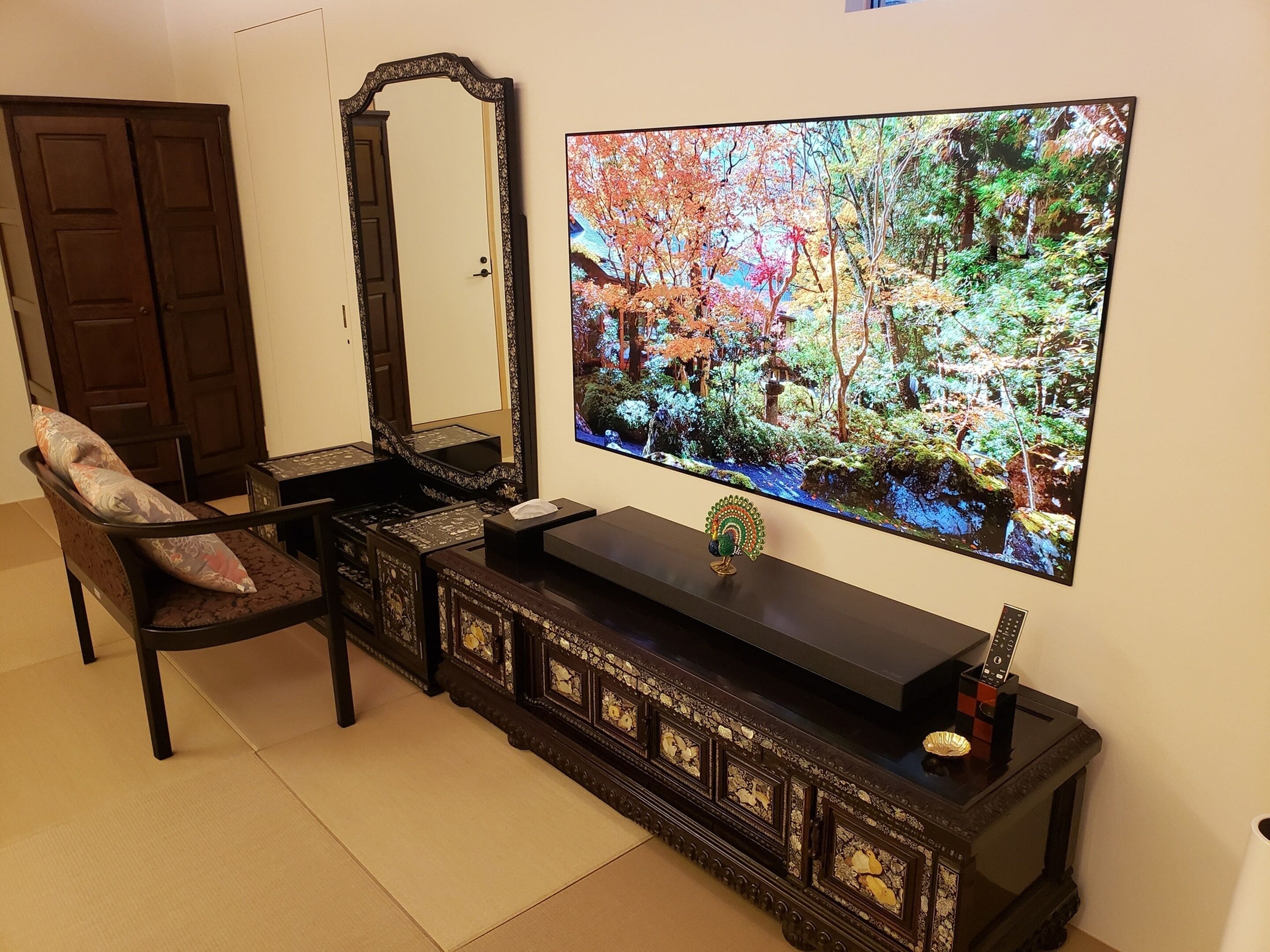 Ein traditioneller japanischer Raum mit Tatami-Böden, einem tiefen, aus Holz gefertigten Schrank mit aufwendigen Schnitzereien, einem großen Flachbildfernseher an der Wand und einer dunkelholzigen Tür.