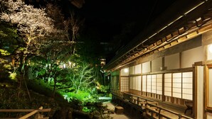 A traditional Japanese garden at night with a wooden walkway, a koi pond, and a building with sliding doors.