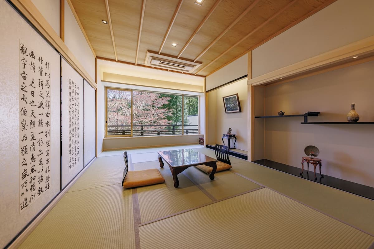 A traditional Japanese room with tatami flooring, a low table, cushions, a sliding door, and a large window with a view of cherry blossoms.