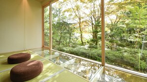 A room with tatami mats, a large window with a view of a garden, and two brown cushions.