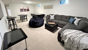 En stue med en grå sofa, en sort skammel, et bordtennisbord og et billardbord. Der er to vinduer og en loftlampe.