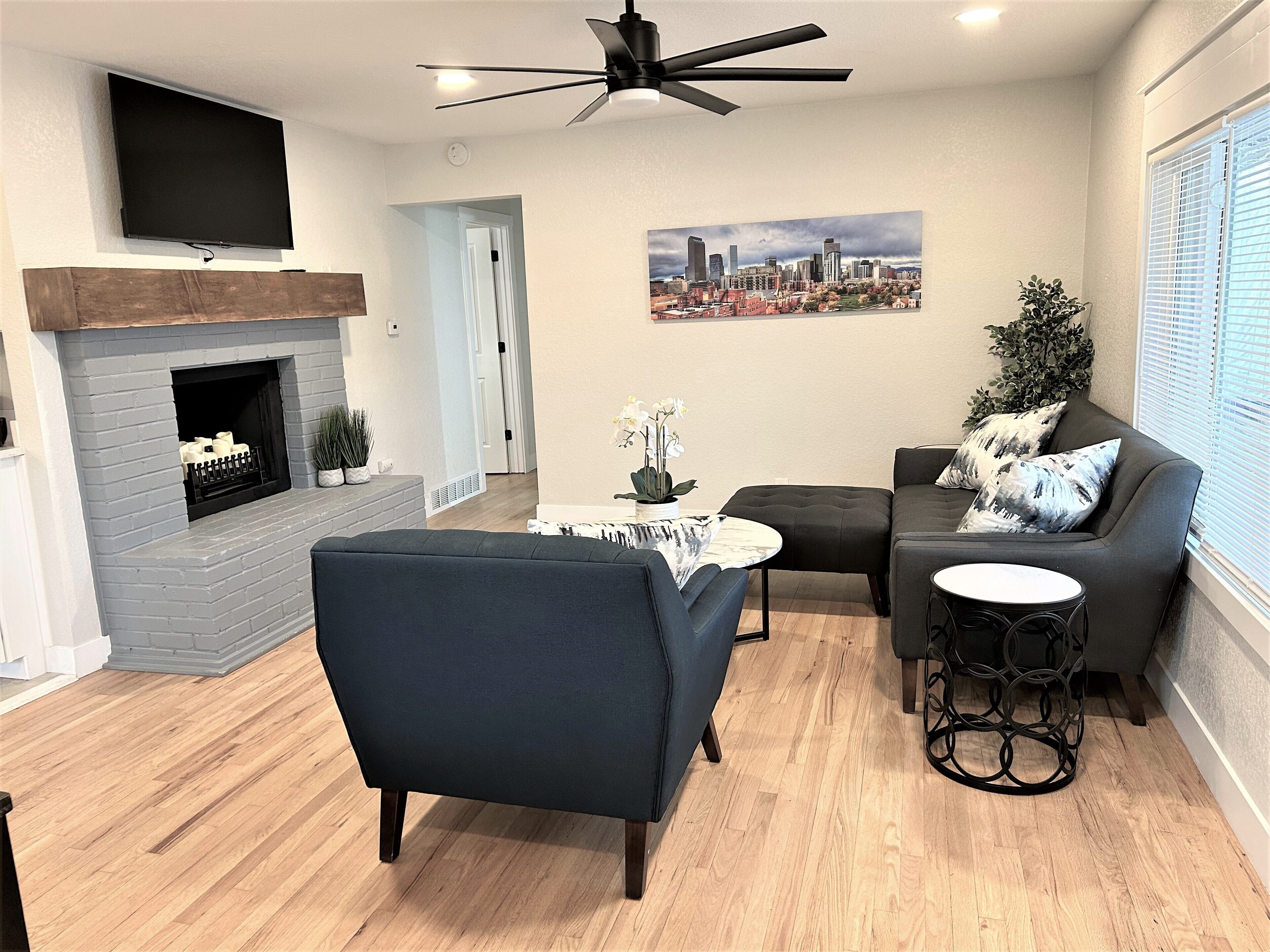 A modern living room with a fireplace, a ceiling fan, a cityscape painting, and a seating area with a couch and chairs.