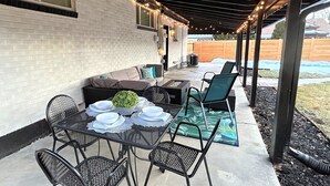 En udendørs terrasse med et bord dækket til fire personer, omgivet af stole, samt et overdækket siddeområde med en pejs.