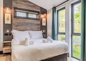 A modern bedroom with a large bed, bedside tables, a built-in bench, a window with curtains, and wall-mounted lighting. - Dovedale Premier Lodge (Ashbourne)