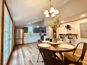 Una cocina moderna con electrodomésticos de acero inoxidable, un comedor con mesa y sillas de madera, y una lámpara colgante.