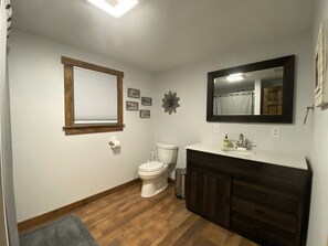 Une salle de bain équipée de toilettes, d’un lavabo, d’un miroir et d’une fenêtre avec des stores.