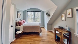 A bedroom with a sloped ceiling, a bed with a beige bedspread, a small bedside table, and a window with a view of trees.