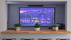 Una habitación moderna con una pantalla grande que muestra un paisaje urbano, dos plantas en macetas sobre una mesa de madera, y una zona de estar con sillas.