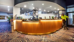 A hotel reception area with a curved wooden reception desk, a digital check-in station, and a patterned carpet. - Nightcap at Empire Hotel (Kilburn)