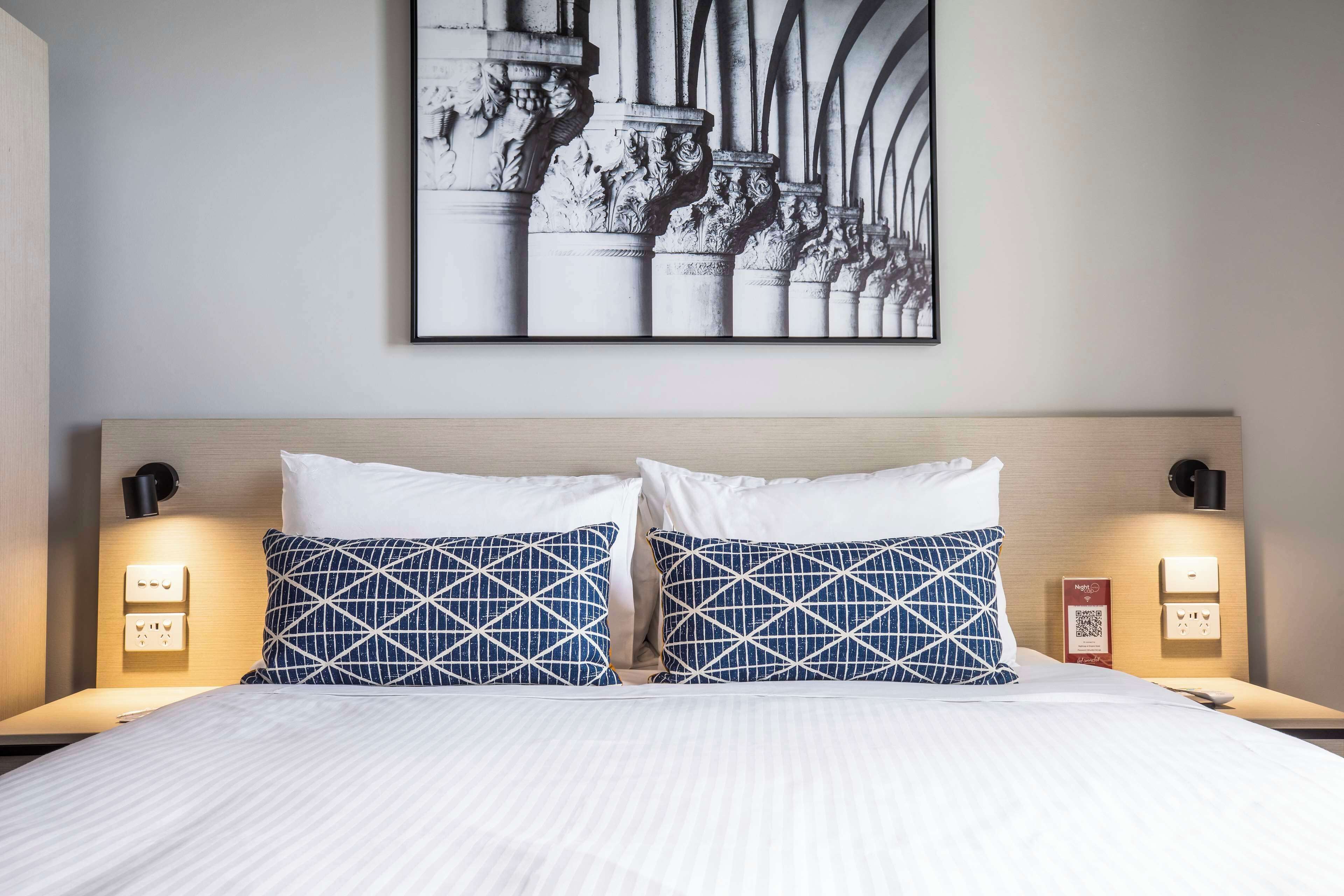 A hotel room with a large bed, two matching blue and white patterned pillows, a framed artwork above the bed, and bedside lamps.