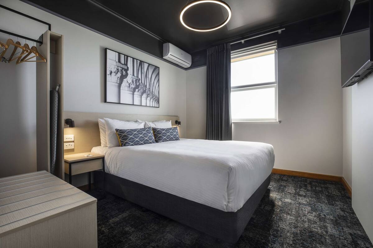Una habitación de hotel moderna con una cama grande, una mesita de noche, un armario y una ventana con cortinas.