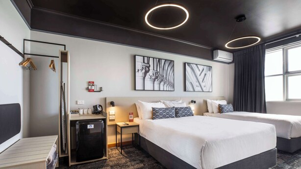 A hotel room with two beds, a desk, a refrigerator, and artwork on the wall. - Nightcap at Empire Hotel (Kilburn)