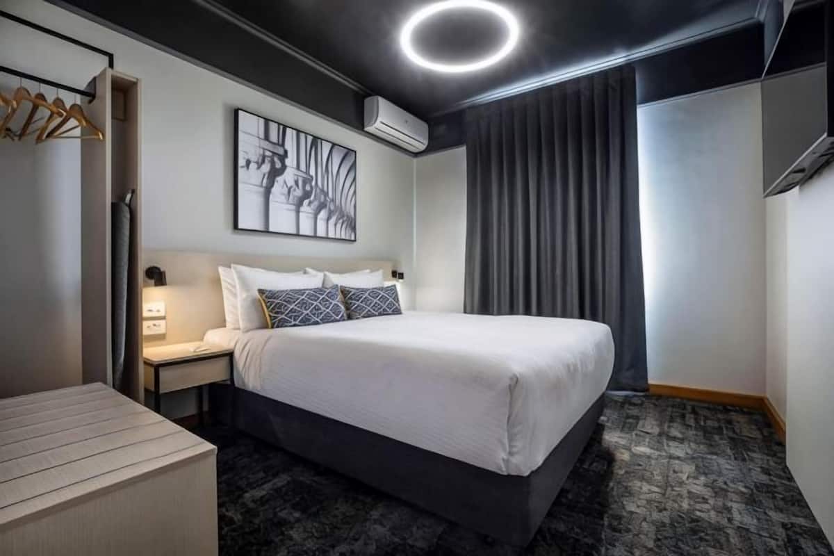 Habitación de hotel con cama, mesitas de noche, escritorio, silla, perchero y un gran ventanal con cortinas.