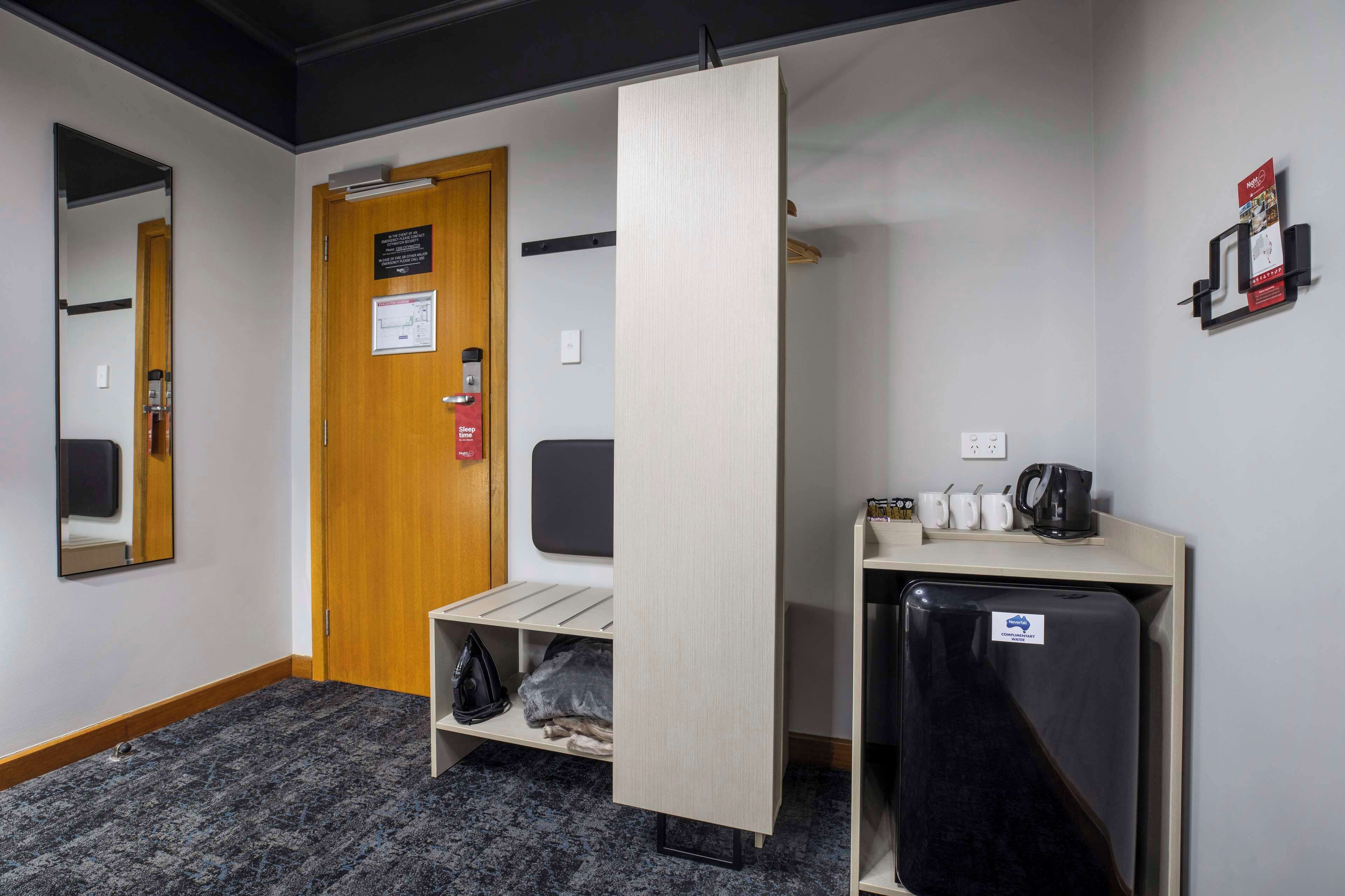 Une chambre d’hôtel avec une porte en bois, un miroir, une étagère avec une bouilloire, des tasses et un réfrigérateur.
