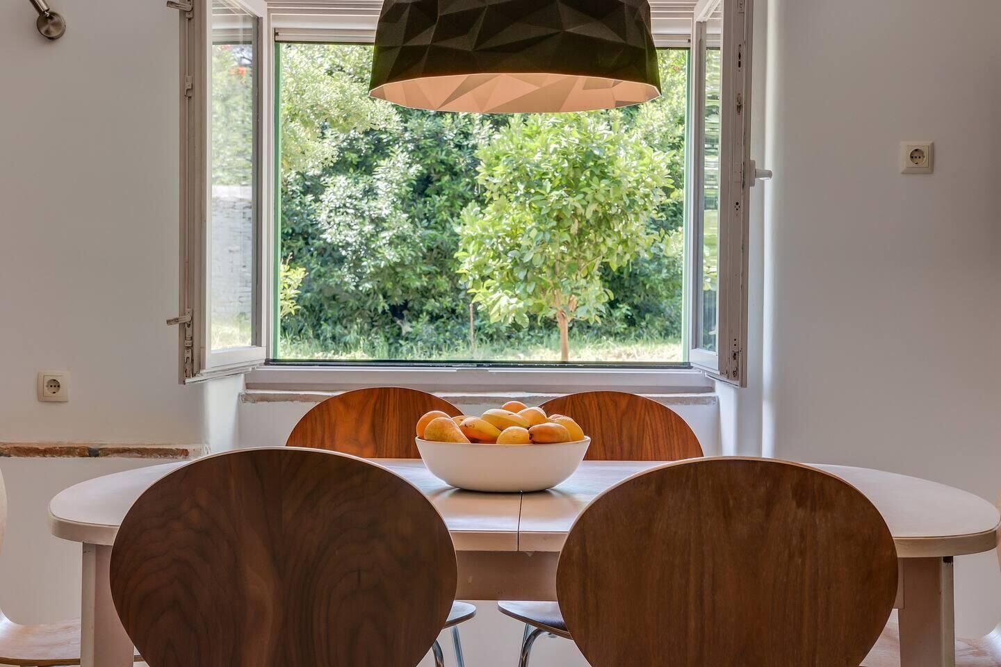 Un coin repas avec une table et des chaises, un saladier de fruits sur la table et une fenêtre donnant sur un paysage verdoyant.