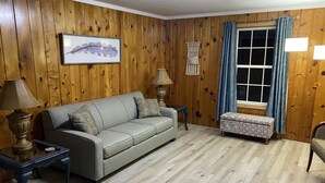 A living room with a wooden paneled wall, a grey sofa, two side tables with lamps, a patterned ottoman, and a window with blue curtains. - Cozy cottage home close to Lake Michigan (Muskegon)