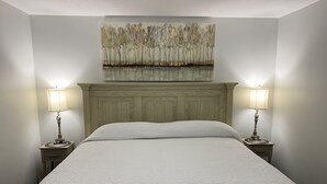 A neatly made bed with a textured white duvet, flanked by two bedside tables with lamps, in a room with a wooden headboard and a painting above the bed. - Cozy cottage home close to Lake Michigan (Muskegon)