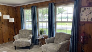 A room with two patterned armchairs, a small round table, a potted plant, and a wall clock. - Cozy cottage home close to Lake Michigan (Muskegon)