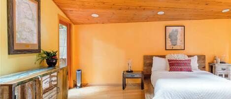 Une chambre avec un plafond en bois, un lit avec une literie blanche et un oreiller rouge, une table de chevet avec une lampe, et une commode en bois ornée d’une plante.