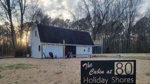 A cabin at Holiday Shores with a dirt ground, a small porch with chairs, and a garage. - Lake Access / Private Boat Launch Cabin with Game Room! (Scottsboro)