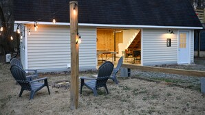 A small, single-story house with a dark roof and white siding. There are two Adirondack chairs outside, a fire pit, and a wooden post with string lights. - Lake Access / Private Boat Launch Cabin with Game Room! (Scottsboro)