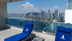 A rooftop terrace with two blue lounge chairs, a glass balustrade, and a view of a city skyline across a body of water. - Best Location, 2 bed, 2baths, High Speed Internet (Panamá)
