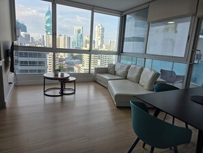 A modern living room with a large window, a sofa, a small round table, and a dining area.