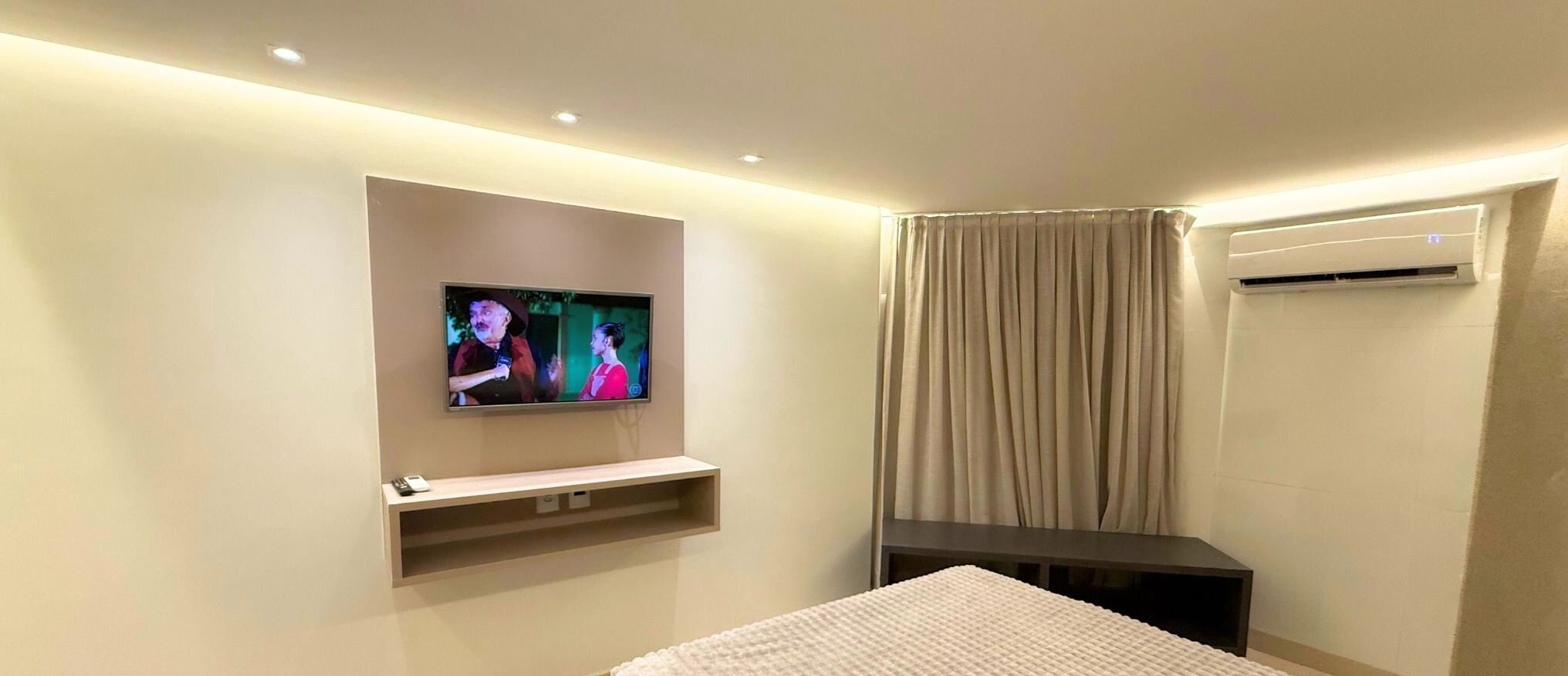 A hotel room with a flat-screen TV mounted on the wall, a bed, a small table, a dark-colored bench, and an air conditioning unit.