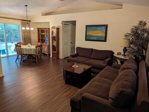 A living room with a brown sofa, a dining table with chairs, a clock, and a painting on the wall.