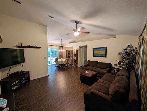 A living room with a flat-screen TV, a ceiling fan, a sofa, a coffee table, and a dining area with a table and chairs.