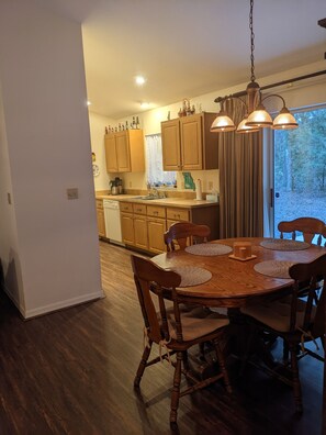 A dining area with a wooden table and chairs, a kitchen in the background with wooden cabinets and a window, and a hanging light fixture.