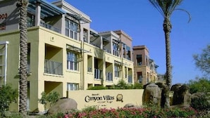 Un hôtel à plusieurs étages doté de balcons, d’une enseigne indiquant « Canyon Villas », et de jardins aménagés avec des palmiers et des rochers.
