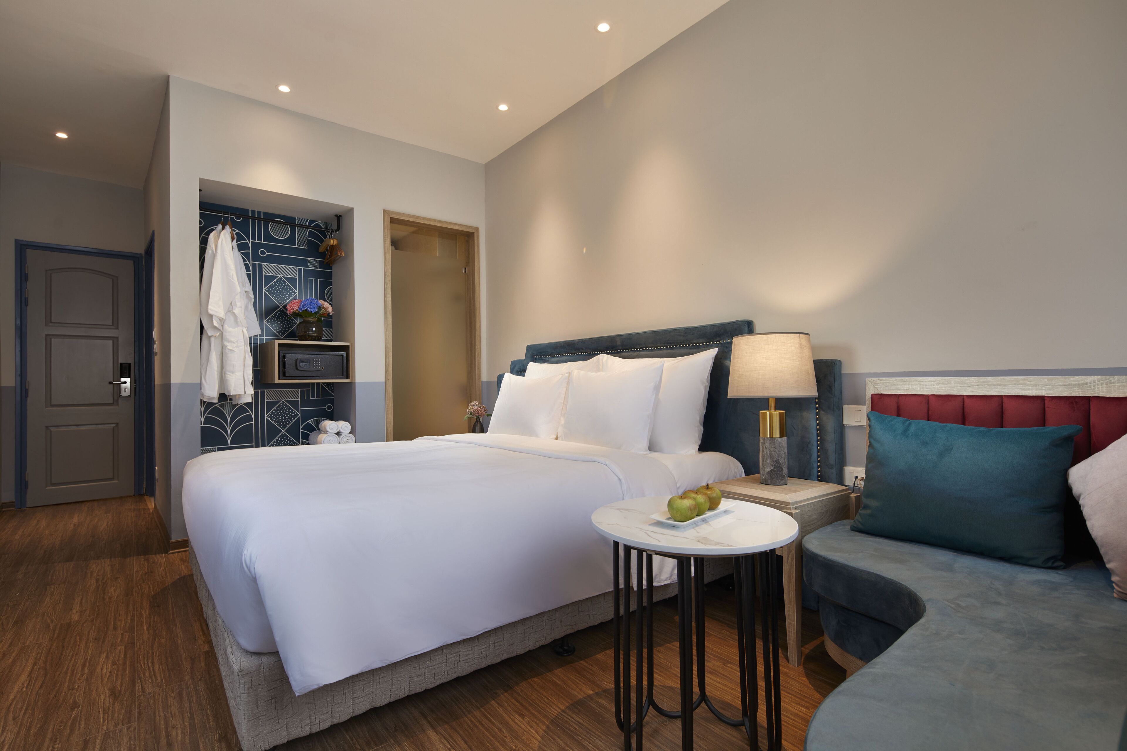Ein Hotelzimmer mit einem großen Bett, einem Nachttisch mit Lampe, einem kleinen runden Tisch mit Obst und einer Sitzecke mit gepolstertem Sitz.