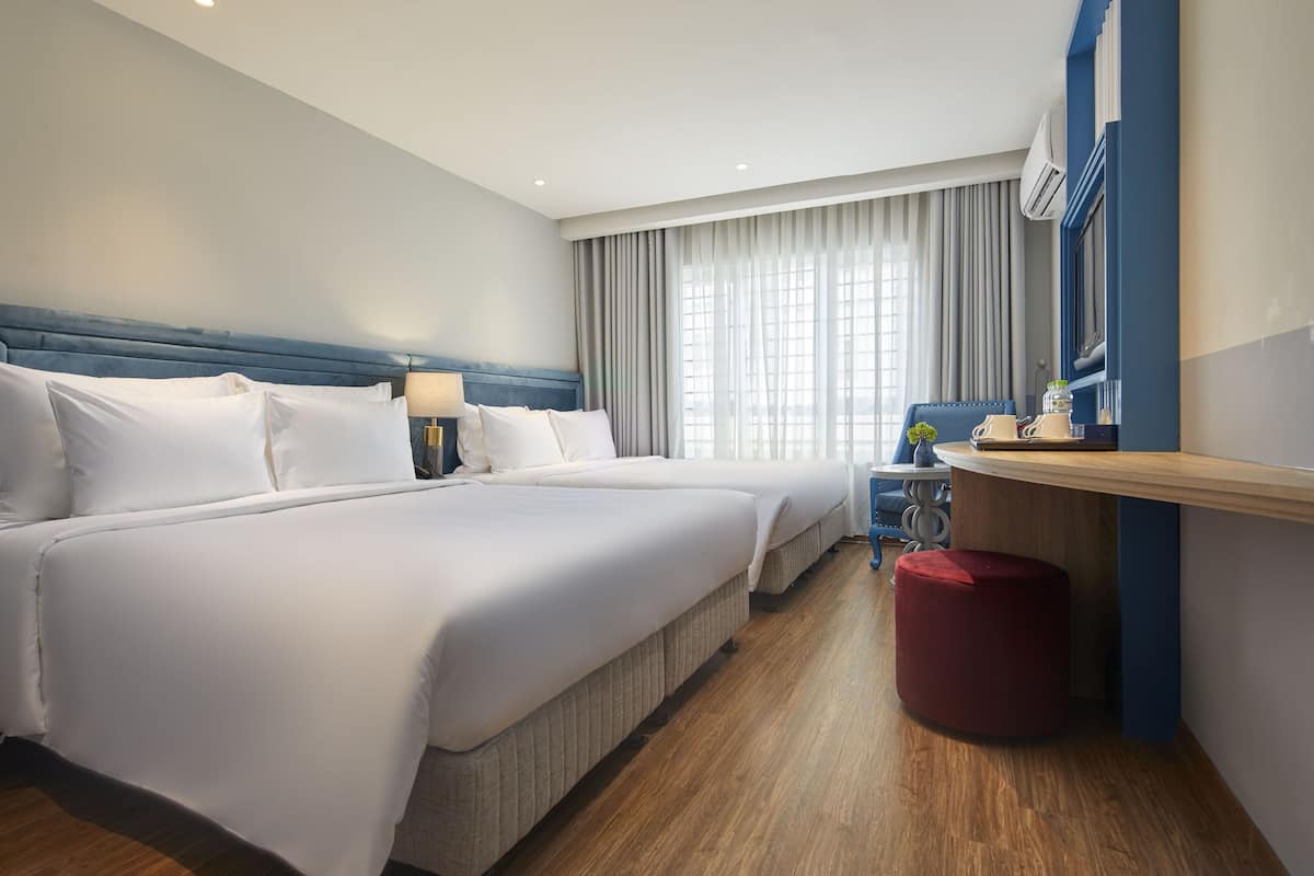 Habitación de hotel con dos camas, un escritorio, un reposapiés rojo y un ventanal con cortinas.