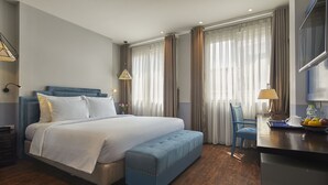 A modern hotel room with a large bed, a blue bench, a desk with a lamp, and a TV.