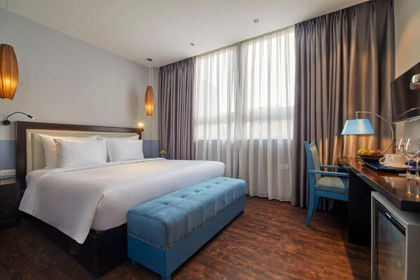 A hotel room with a large bed, a blue bench, a desk with a lamp, and a TV.