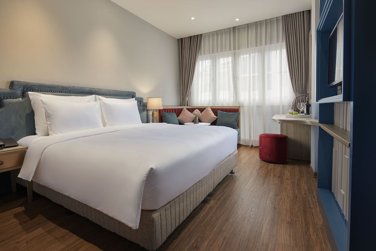 Una habitación de hotel con una cama grande, una mesita de noche, un reposapiés rojo, un televisor montado en la pared y una ventana con cortinas.