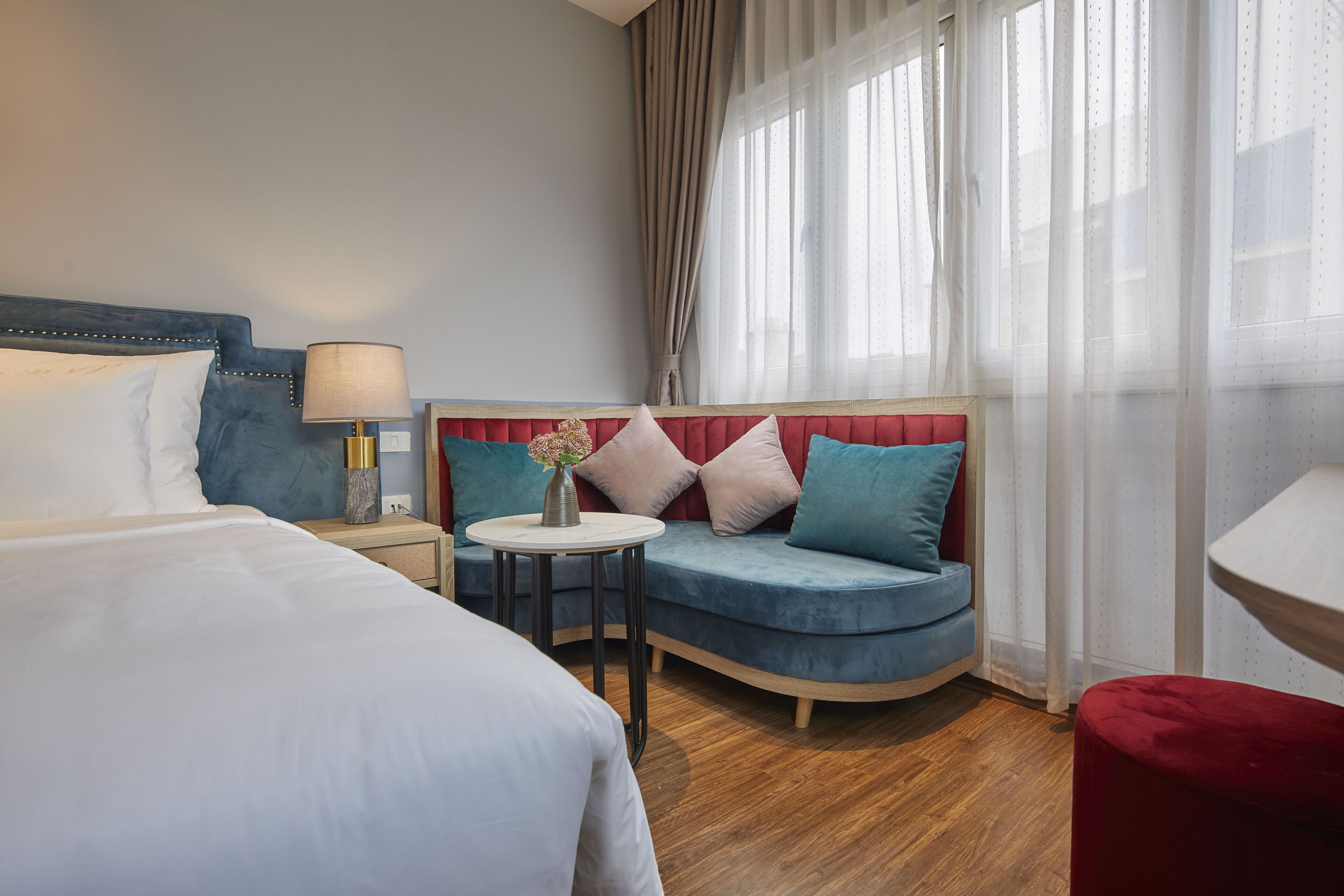 Una habitación de hotel con una cama, un sofá, una mesita con un florero y un reposapiés rojo.