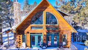 A log cabin with a stone chimney, large windows, and a porch with chairs. - LOG CABIN w/Mtn View, Hot Tub, & Fireplace! (Divide)