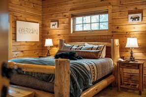 A log cabin bedroom with a bed, two bedside lamps, a window, and wall art. - LOG CABIN w/Mtn View, Hot Tub, & Fireplace! (Divide)