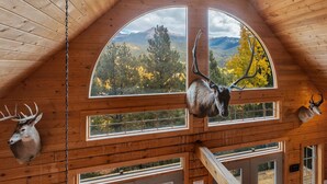 A log cabin with a high ceiling, wooden beams, and multiple windows offering views of a mountainous landscape. - LOG CABIN w/Mtn View, Hot Tub, & Fireplace! (Divide)