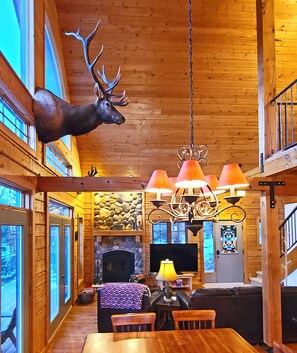 A log cabin interior with a large deer head mounted on the wall, a fireplace, a television, and a dining area with a table and chairs.