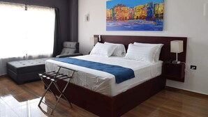 A modern hotel room with a large bed, a grey sofa, a small table, and a painting of colorful buildings on the wall.