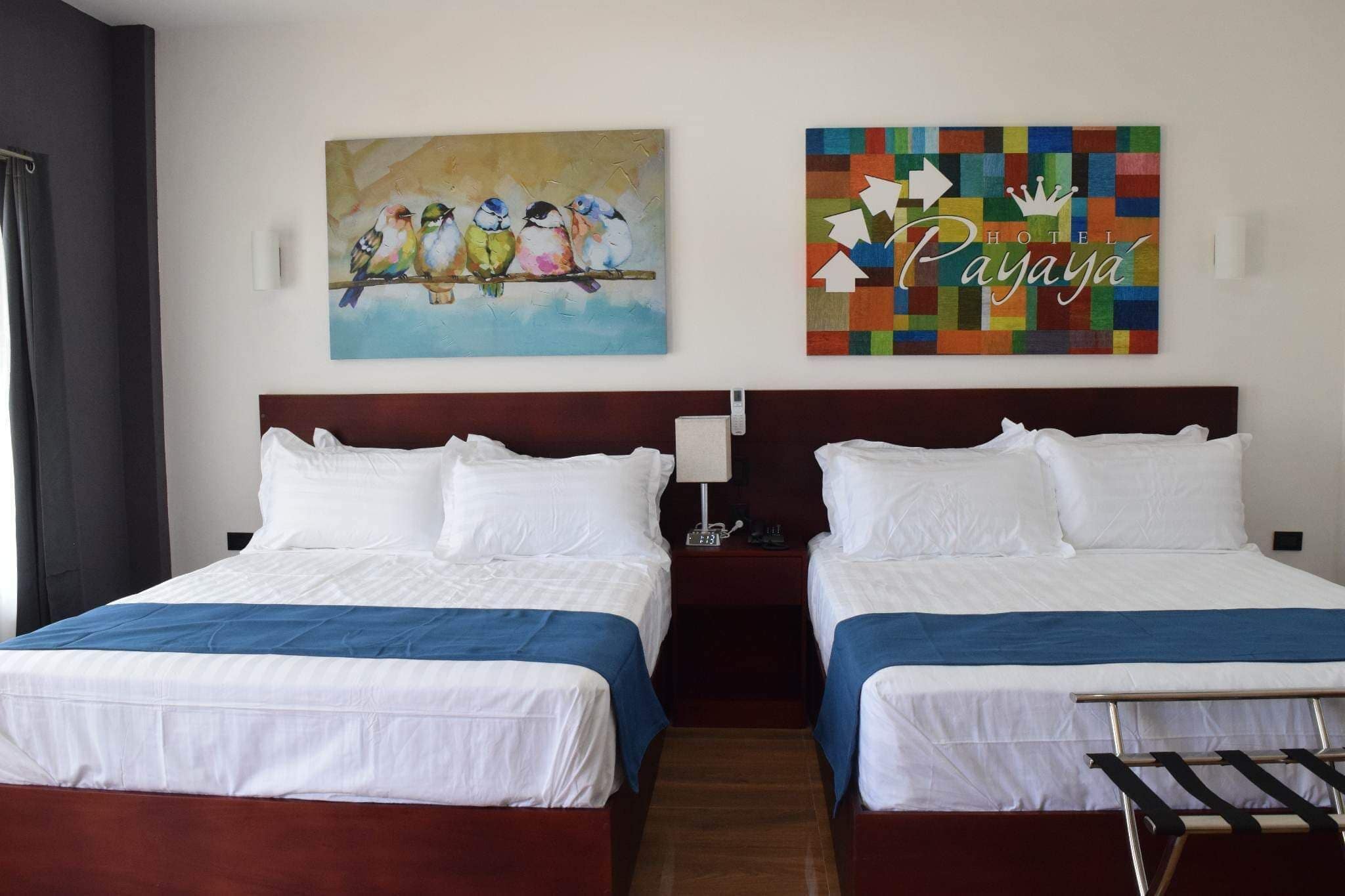 A hotel room with two beds, each with white linens and blue bedspreads, a wooden headboard, and a wall-mounted painting of birds.