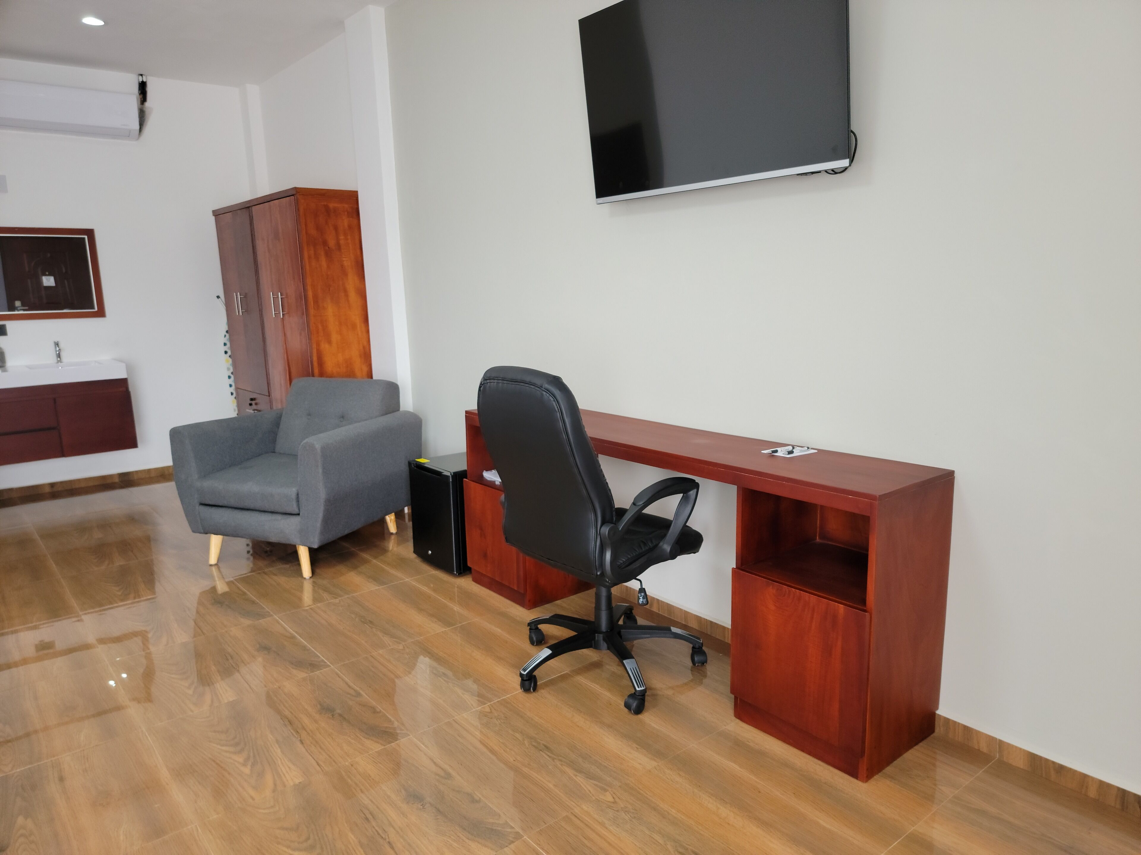 Een moderne kantoorruimte met een houten bureau, een zwarte bureaustoel, een grijze bank en een wandgemonteerde flatscreen-tv.