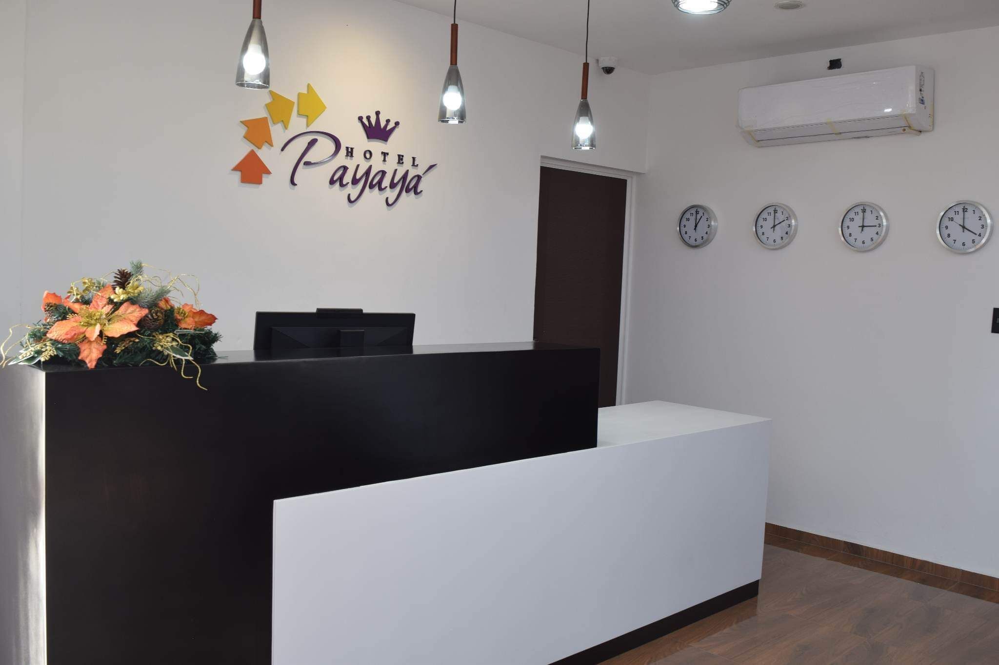 A hotel reception desk with a black counter, a floral arrangement, and a sign that reads "HOTEL Payaya". There are four clocks on the wall and an air conditioning unit above.