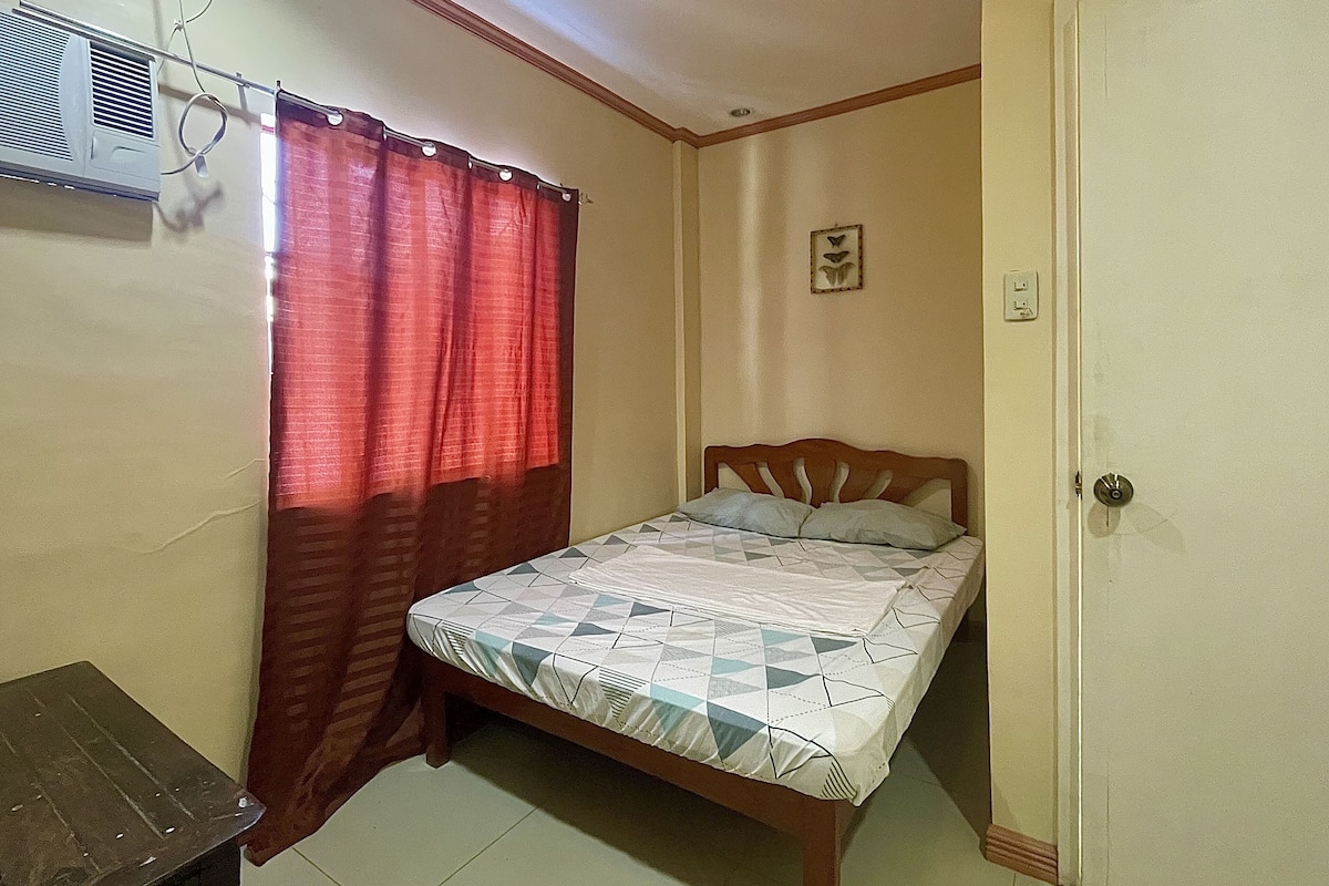 A bedroom with a bed, a wooden headboard, a nightstand, a red curtain, and an air conditioning unit.